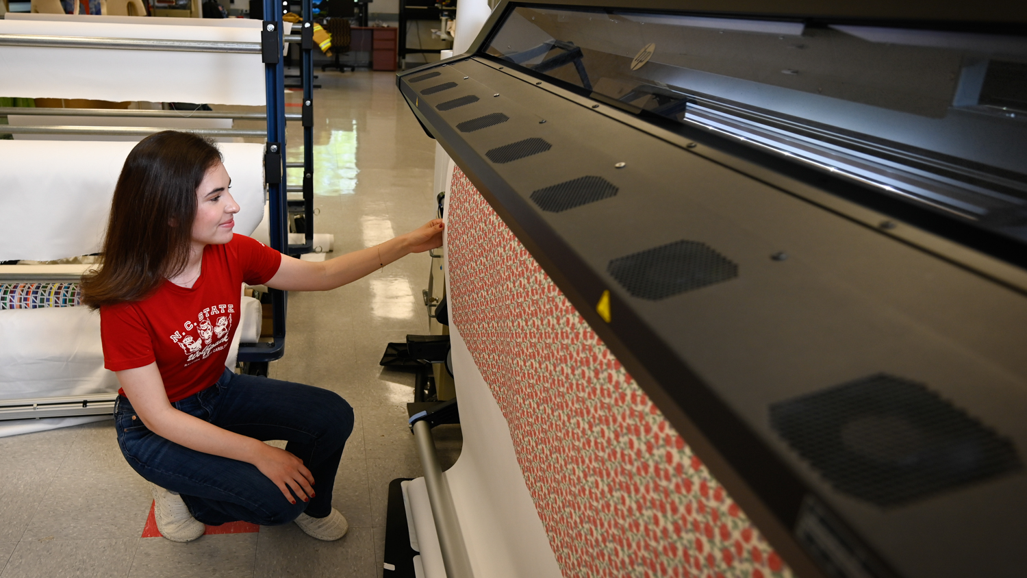 A person wearing a red shirt and blue jeans operates a large printing machine, which is outputting patterned material.