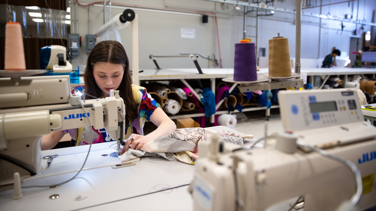 Lindsey Seidenstein operates a sewing machine with a table and gray wall behind her.