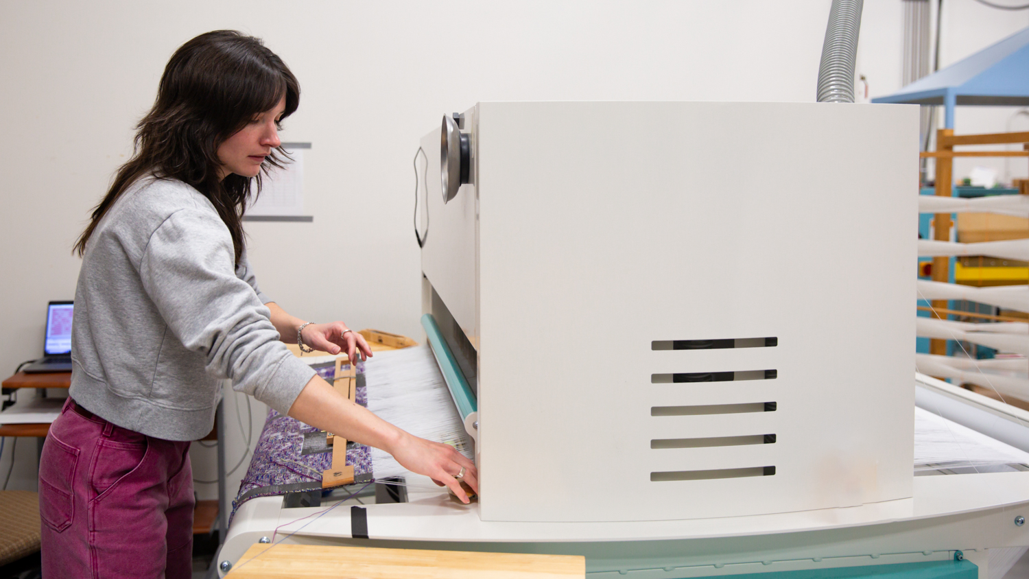 A side view of Chase Woodson operating a TC2 loom. The machinery is white and aqua blue. 