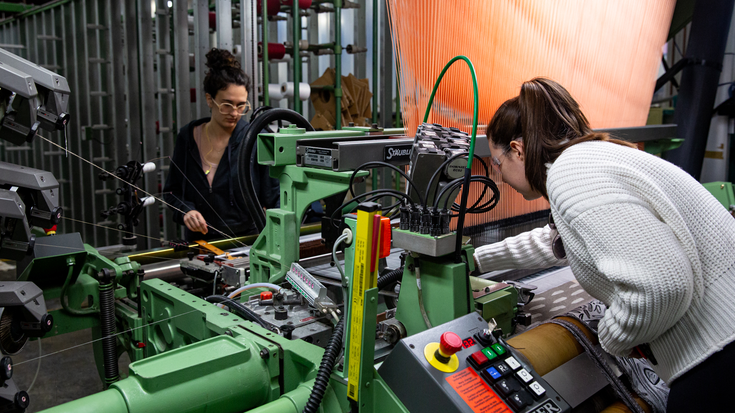 Two people, one wearing glasses and a black outfit, and the other in a white sweater, are operating a Jacquard weaving loom in The Weaving Lab. 