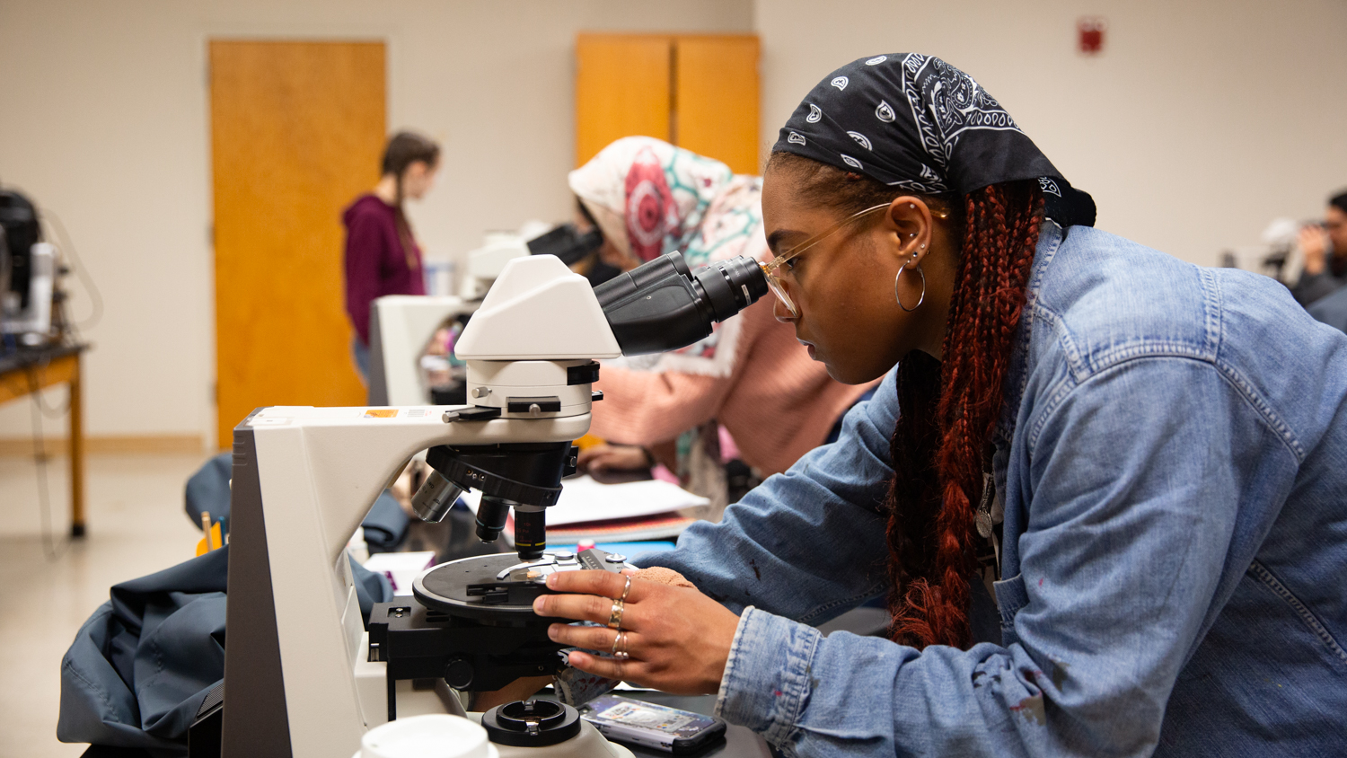 A student in a denim jacket and bandana is looking through a microscope in a classroom. Other students are also engaged in their own work in the background. The room has white walls, large wooden doors, and some lab equipment on the desks.
