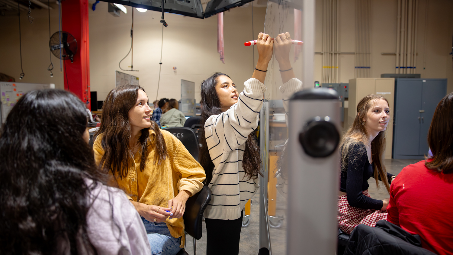 Groups of people are in a Senior Design Lab. One is standing and writing on a dry-erase board with a red marker, while the others are seated and watching her. On the other side of the white board, two other students can be seen talking to each other at a table. 