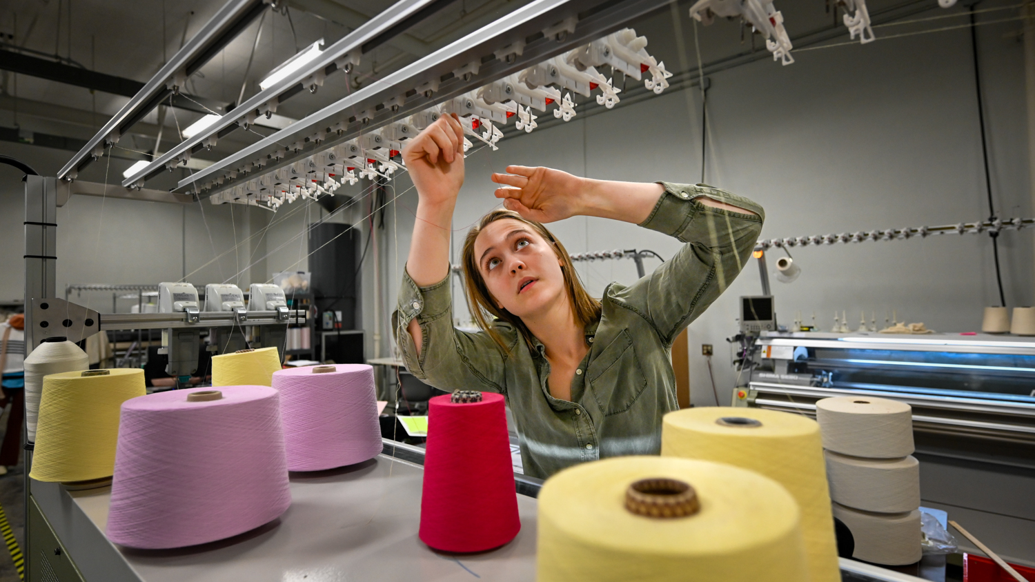 Shayleigh Larsen is adjusting threads in the knitting lab. She is surrounded by large spools of yarn in various colors, including pink, yellow, and white. The environment is a modern, well-lit workspace with knitting machinery in the background.