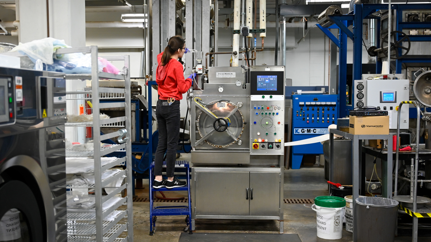 Sophie Frain is wearing a red hoodie and stands on a blue step stool while operating a large industrial machine with multiple buttons and dials in The Dyeing and Finishing Lab. 